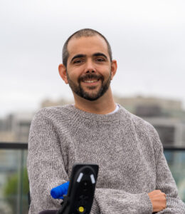 Alex Ghenis, disability and climate change consultant. White man with dark features, close cut hair and beard, smiling. Gray sweater. Low buildings in background. Wheelchair joystick in foreground.