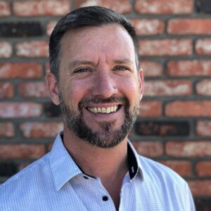 Professional profile photo. White man, beard, blue button up shirt, brick background
