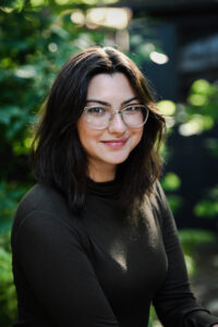 Professional profile photo, white woman, brown hair, glasses, dark shirt. Plants in the background