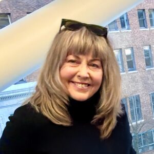 Profile photo, white woman smiling, very light brown mid-length hair, black shirt, brick building in background
