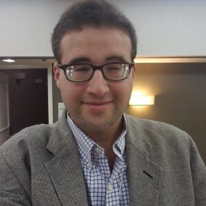 Profile photo of a Caucasian man with short brown hair, black rimmed glasses, a blue-and-white button up shirt and a gray blazer